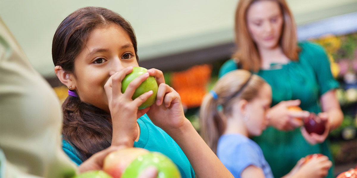 A child eats a green apple while a mother and child pick out red apples in the background