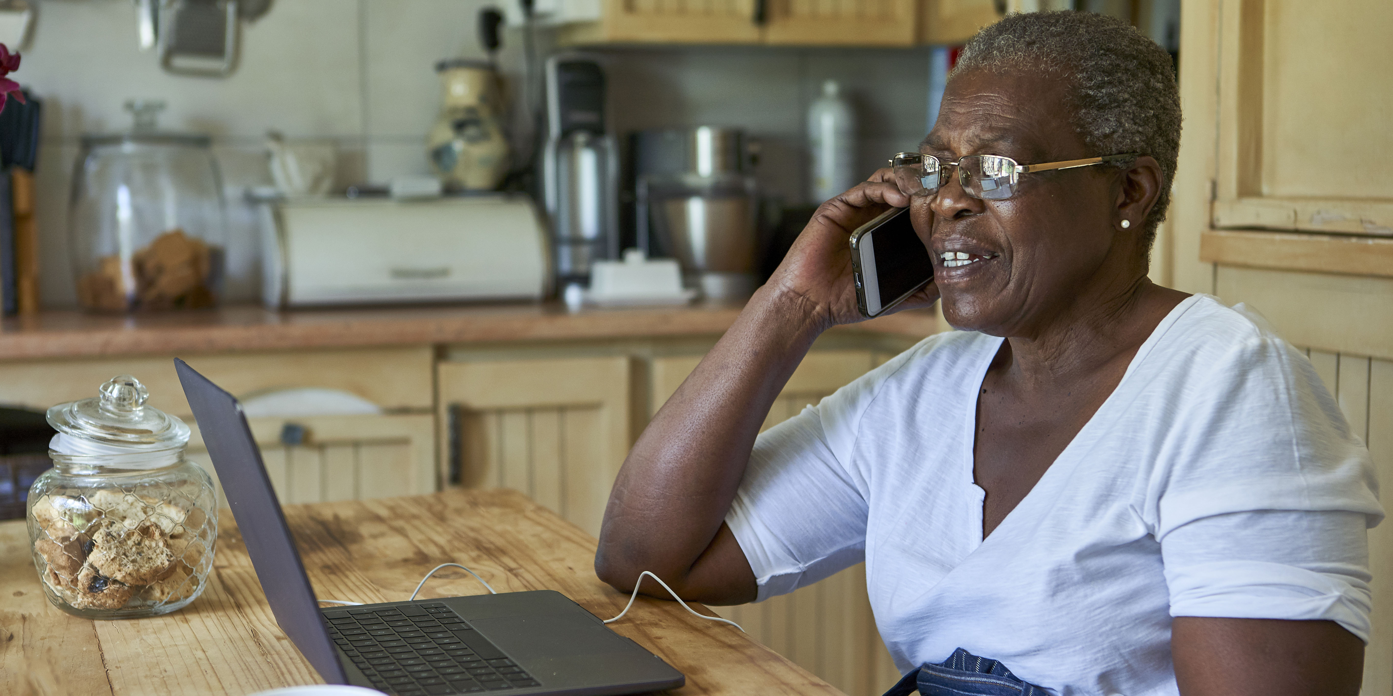 Senior woman sitting at kitchen table using laptop and smartphone